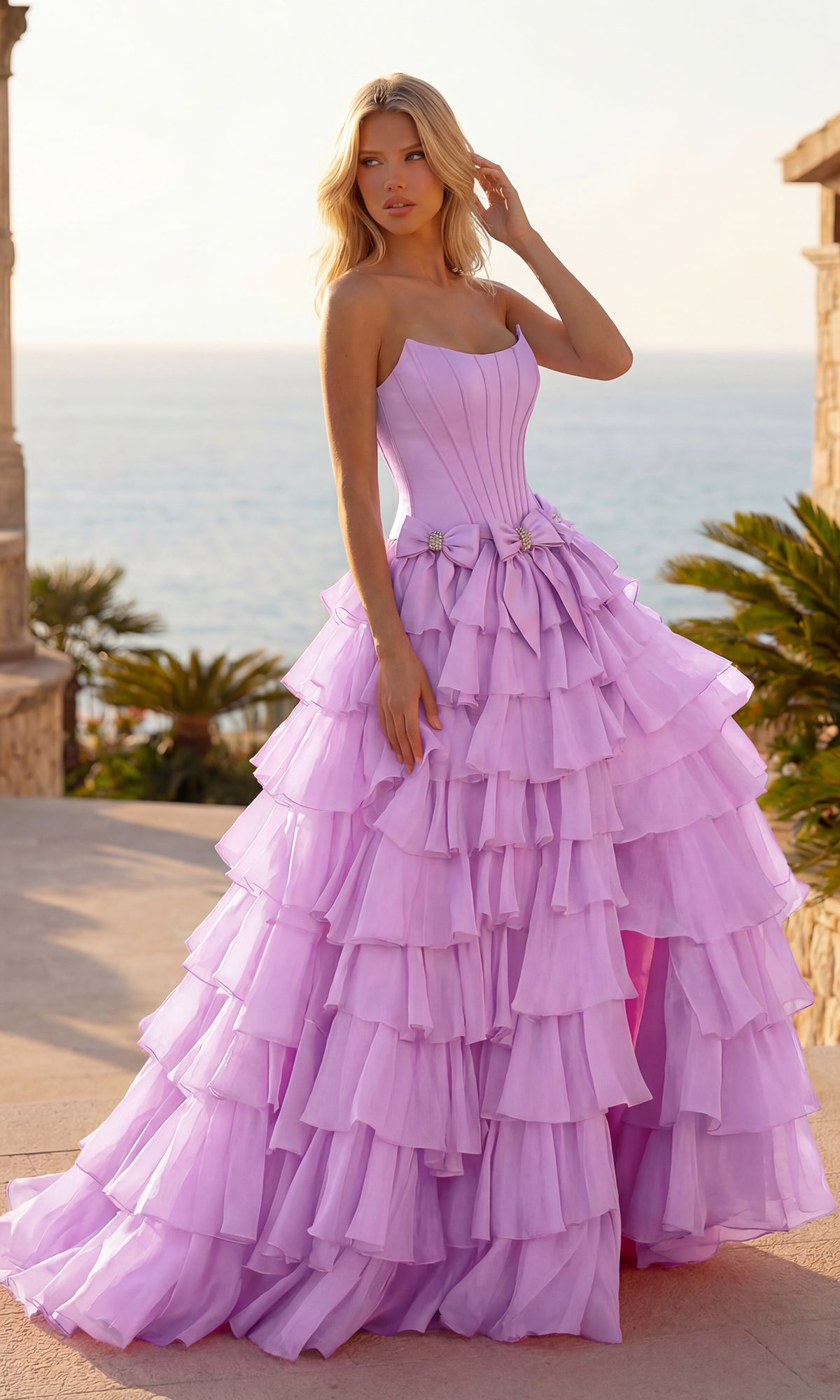 A woman in a lilac Tiered Prom Ball Gown with Bows (PromGirl 250071) stands outdoors near a stone structure with the ocean in the background.
